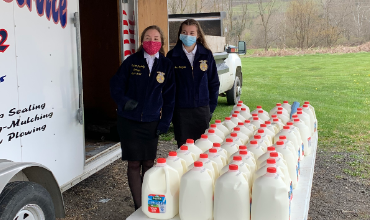 Two students in masks stand behind table with gallons of milk jugs